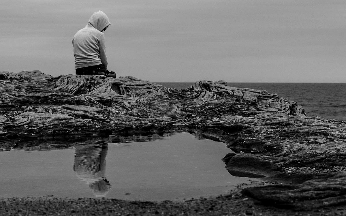 sedící člověk na břehu, kapuce, schoulení smutek / Photo by Clément Falize on Unsplash
