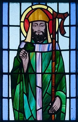 A stained glass window depicts Saint Patrick dressed in a green robe with a halo about his head, holding a sham rock in his right hand and a staff in his left.
Saint Patrick depicted in a stained-glass window at Saint Benin's Church in Kilbennen, near Tuam, in County Galway, Ireland / Wikimedia
